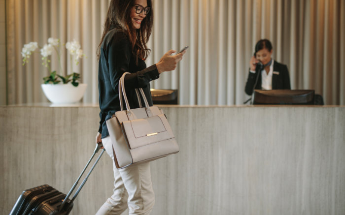 En glad kvinna med glasögon, en stor handväska och en rullväska går i en hotelllobby medan en receptionist pratar i telefon i bakgrunden